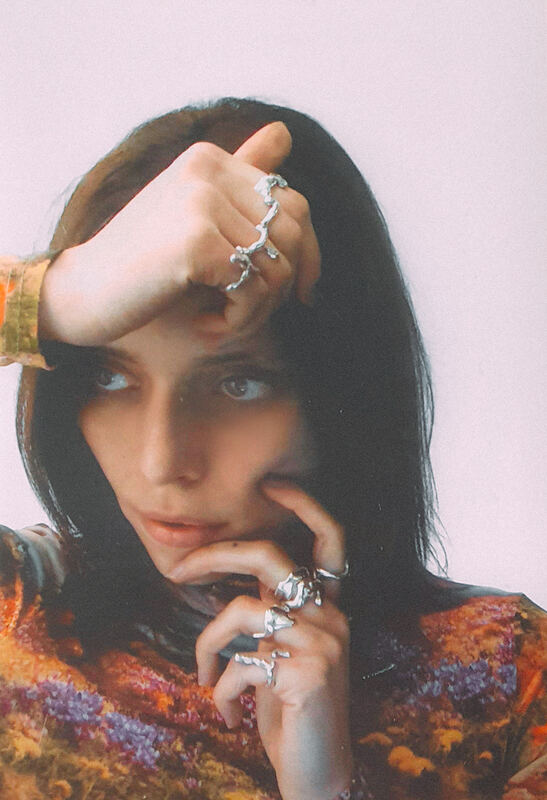 Fanny with a hand on her forehead, rings stacked on several fingers, in a painted floral top